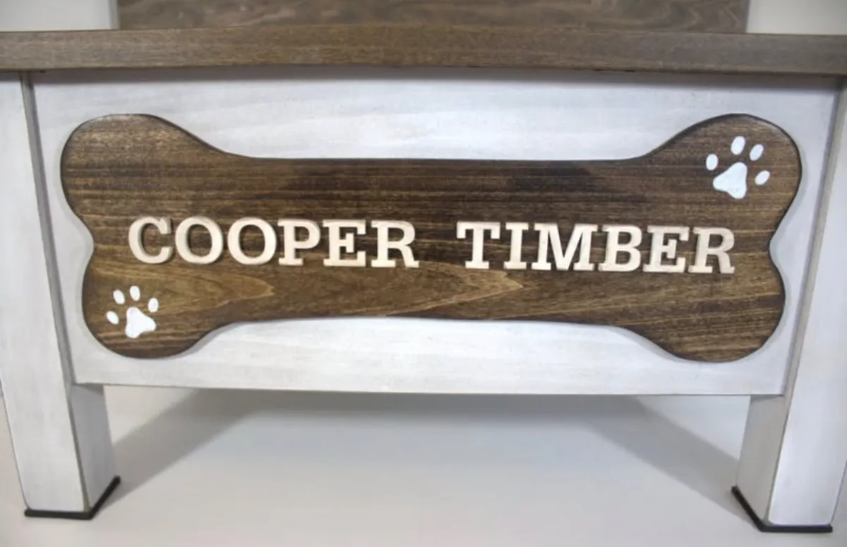 Close-up of a dark stained dog bone nameplate with white wood letters spelling Cooper Timber and two painted white paw prints
